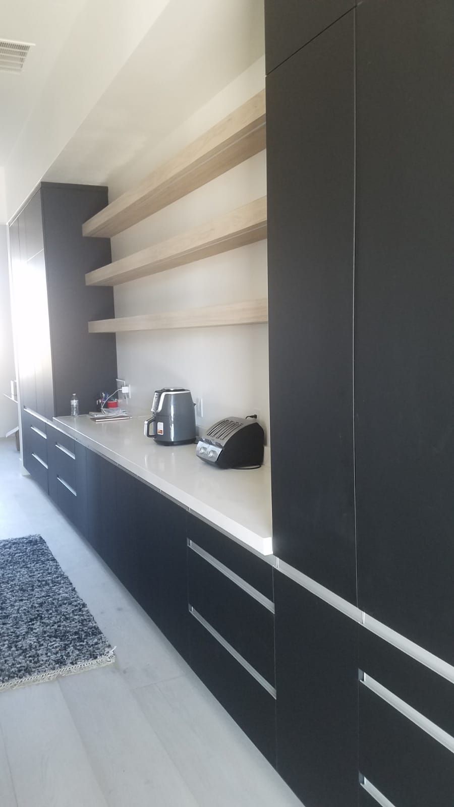 Modern kitchen with gray cabinets, light shelves, and countertop; appliances and rug present.