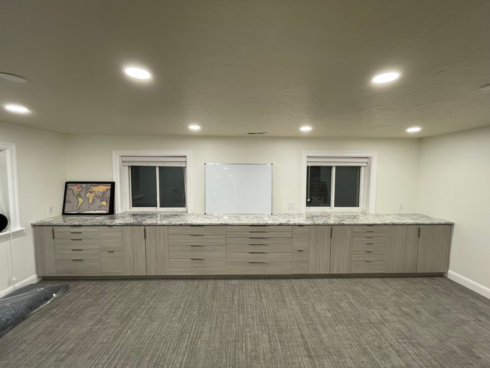 A long, light-colored cabinet with drawers and countertop, below two windows, white board, and recessed ceiling lights.