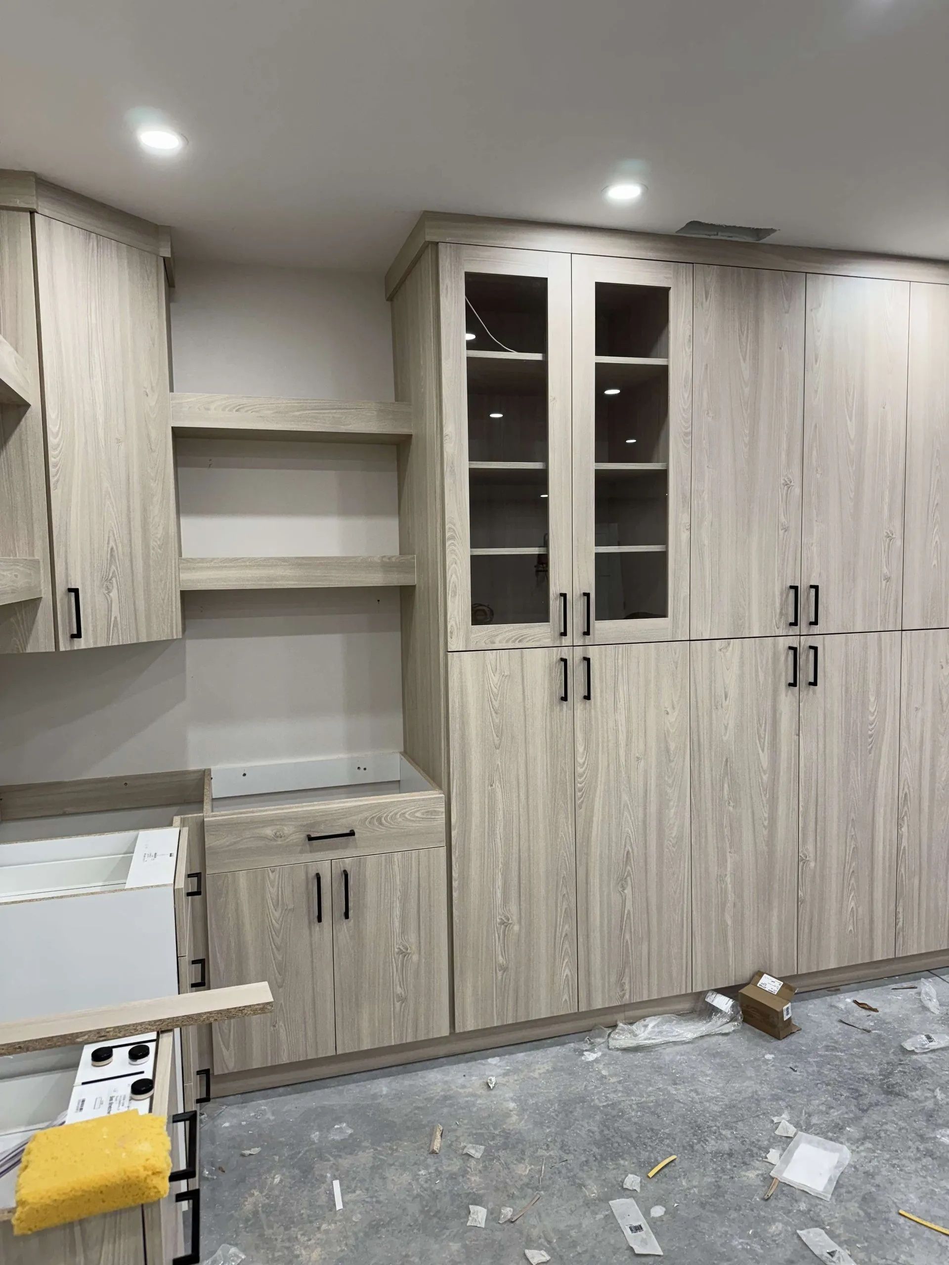 Light wood-grain kitchen cabinets, shelves, and glass-door pantry units in a newly renovated space.