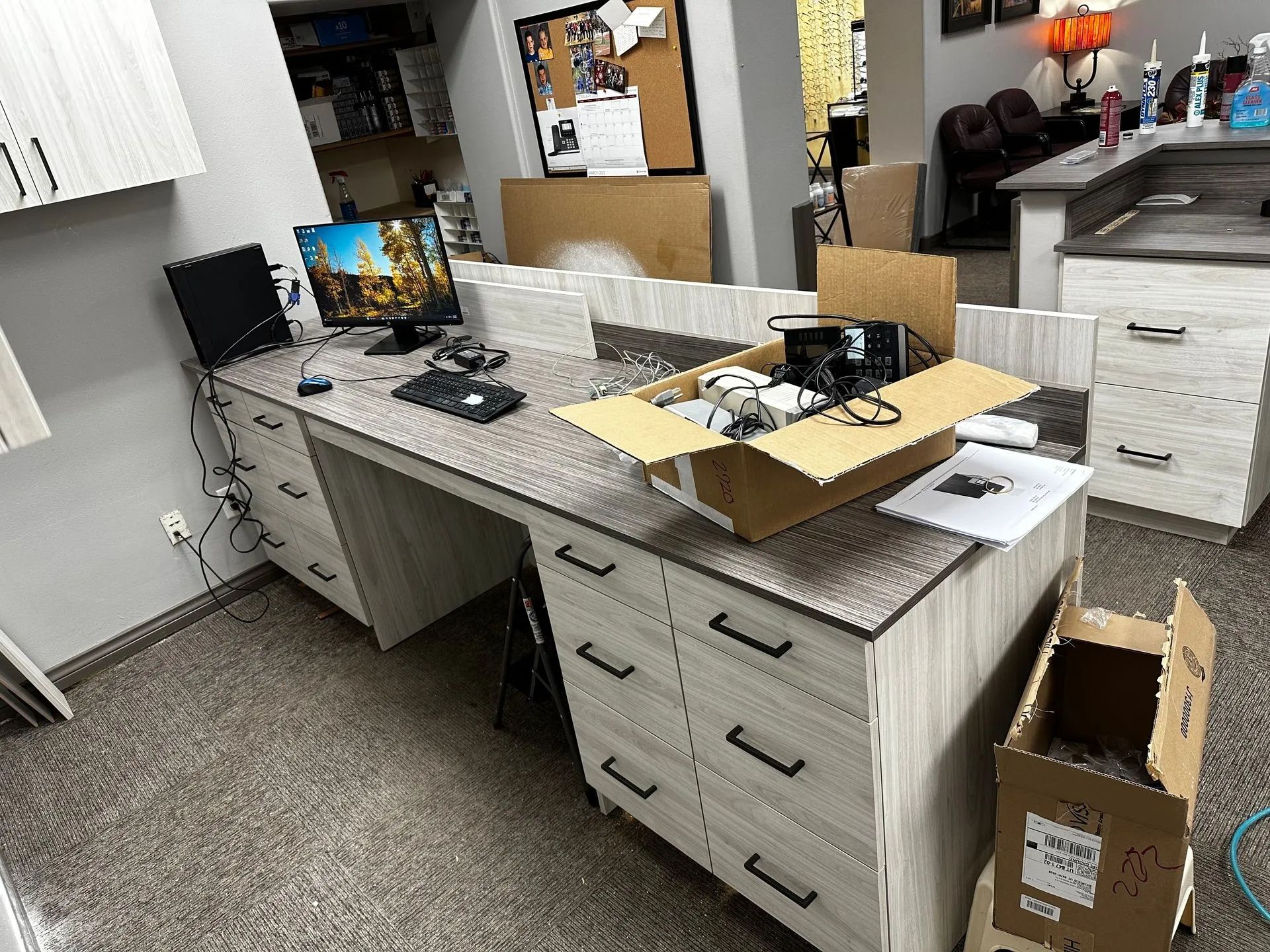 Office desk with computer, drawers, and open cardboard box. Light-colored wood finish, gray countertop, indoor setting.