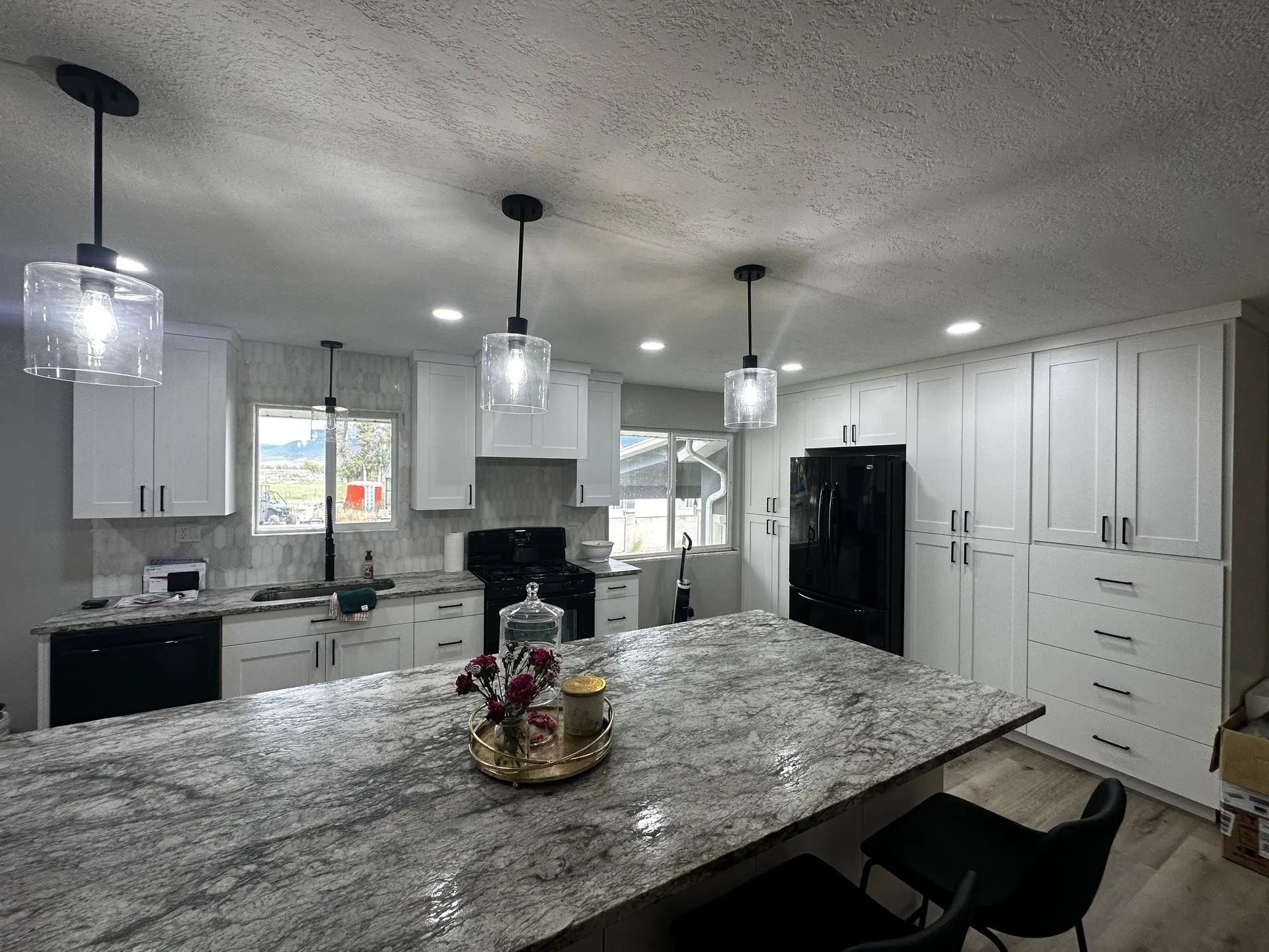 Modern kitchen with white cabinets, large granite island, pendant lights, and black appliances.