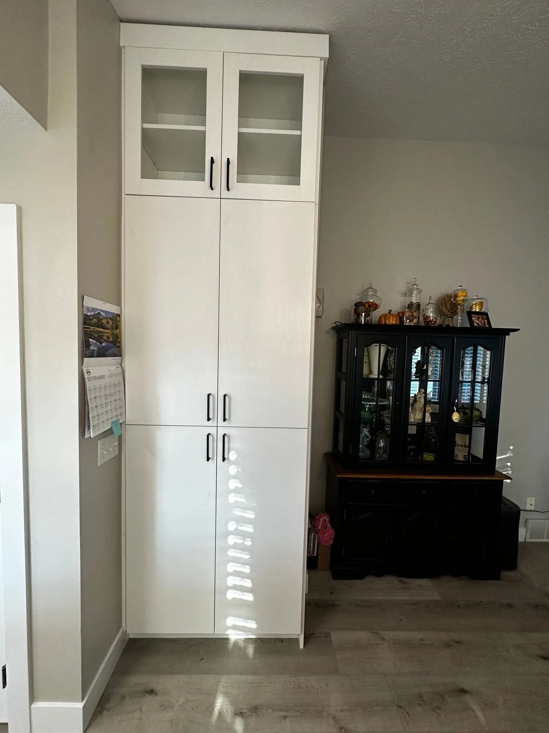 Tall white pantry cabinet with glass-front upper doors, next to a black cabinet on light wood flooring.