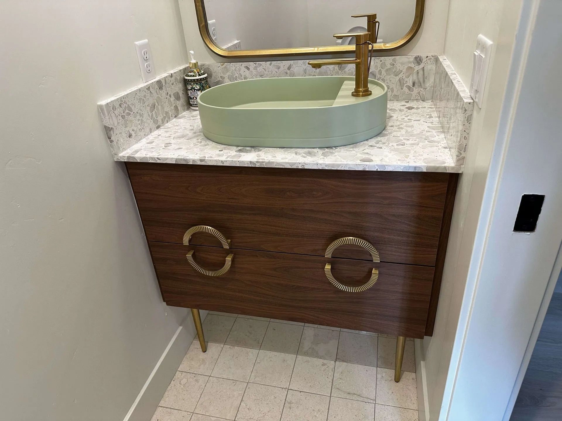 Bathroom vanity with green vessel sink, gold faucet, and decorative pulls, brown cabinet with gold legs.