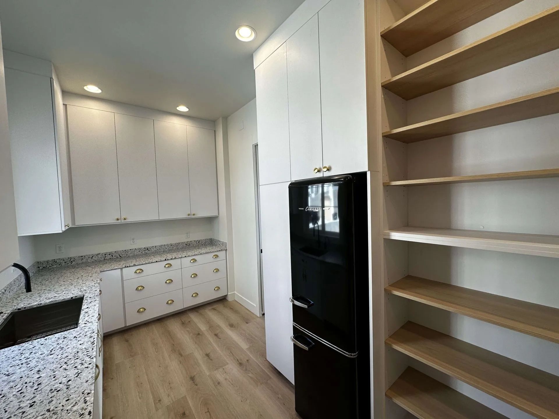 White kitchen with black refrigerator and pantry shelves; speckled countertops, wood flooring.