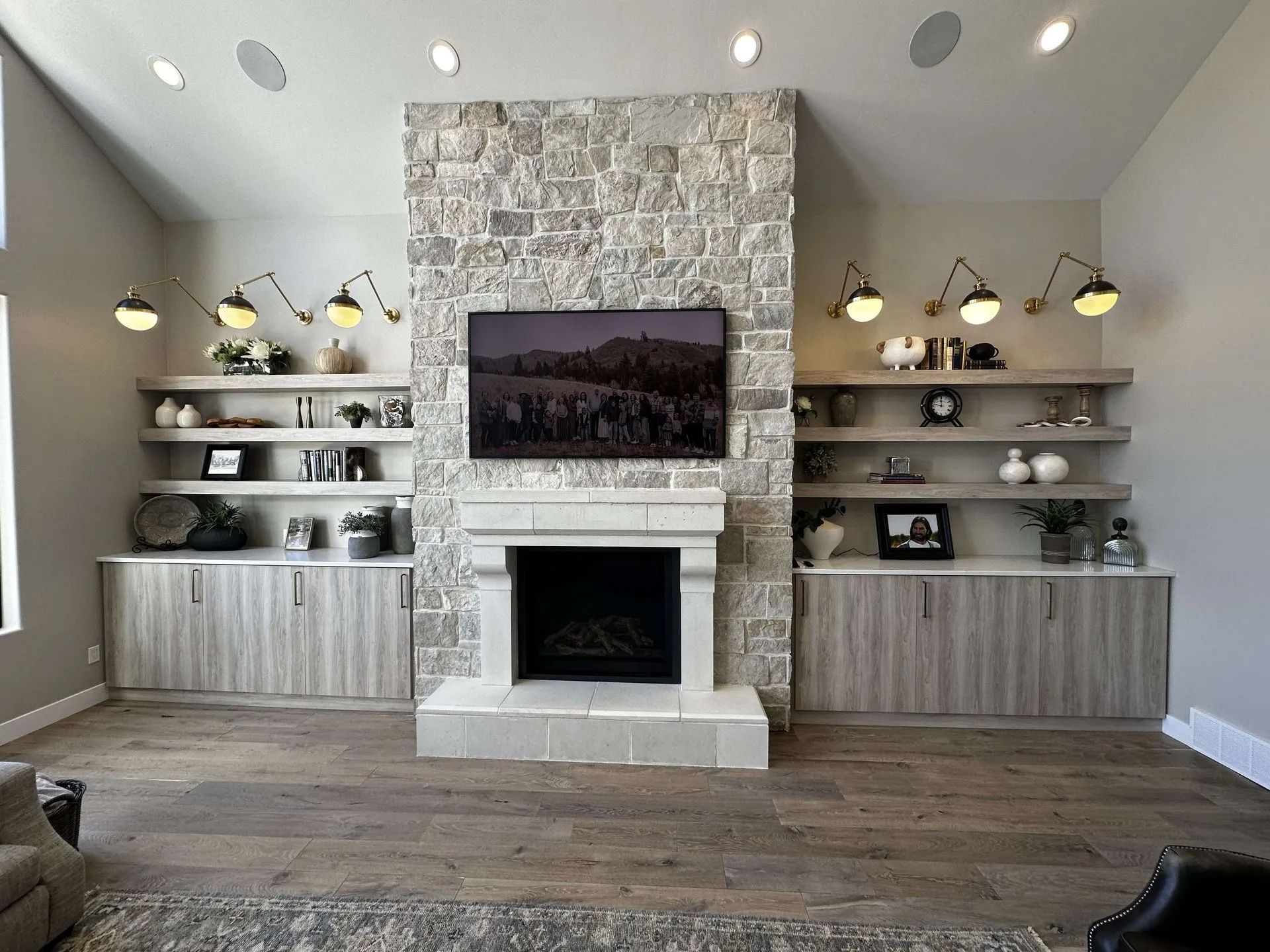Living room with stone fireplace, built-in shelves, TV, and cabinets, featuring warm lighting and wood flooring.