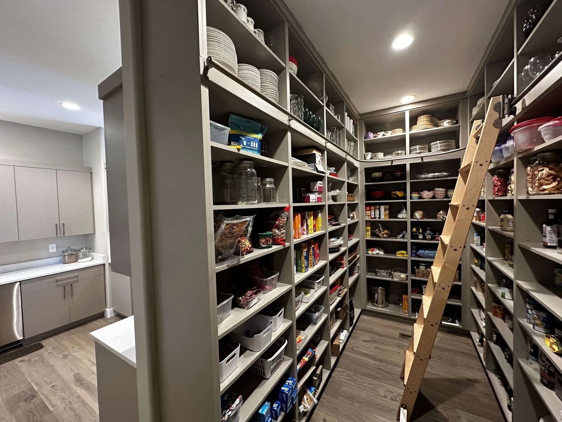 A walk-in pantry with gray shelves full of food items and a wooden ladder. Adjacent is a small kitchen area.