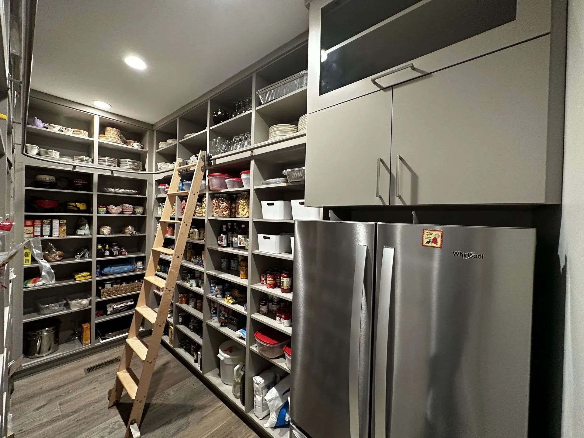 Pantry with shelving and ladder, alongside a refrigerator, all in a room with hardwood flooring.