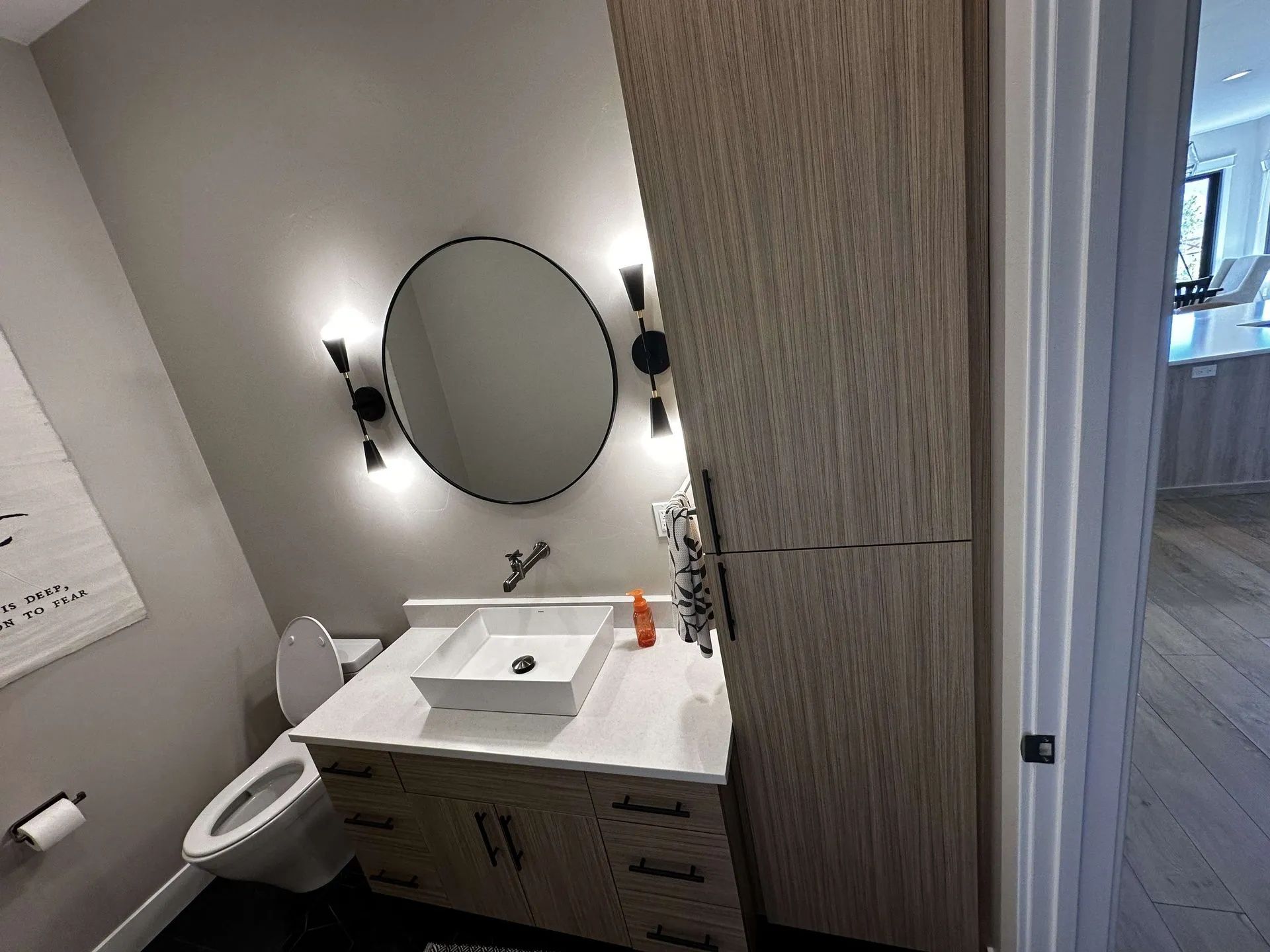 Bathroom with oval mirror, cabinet, and white square sink. Wooden cabinet on the right. Toilet on the left.