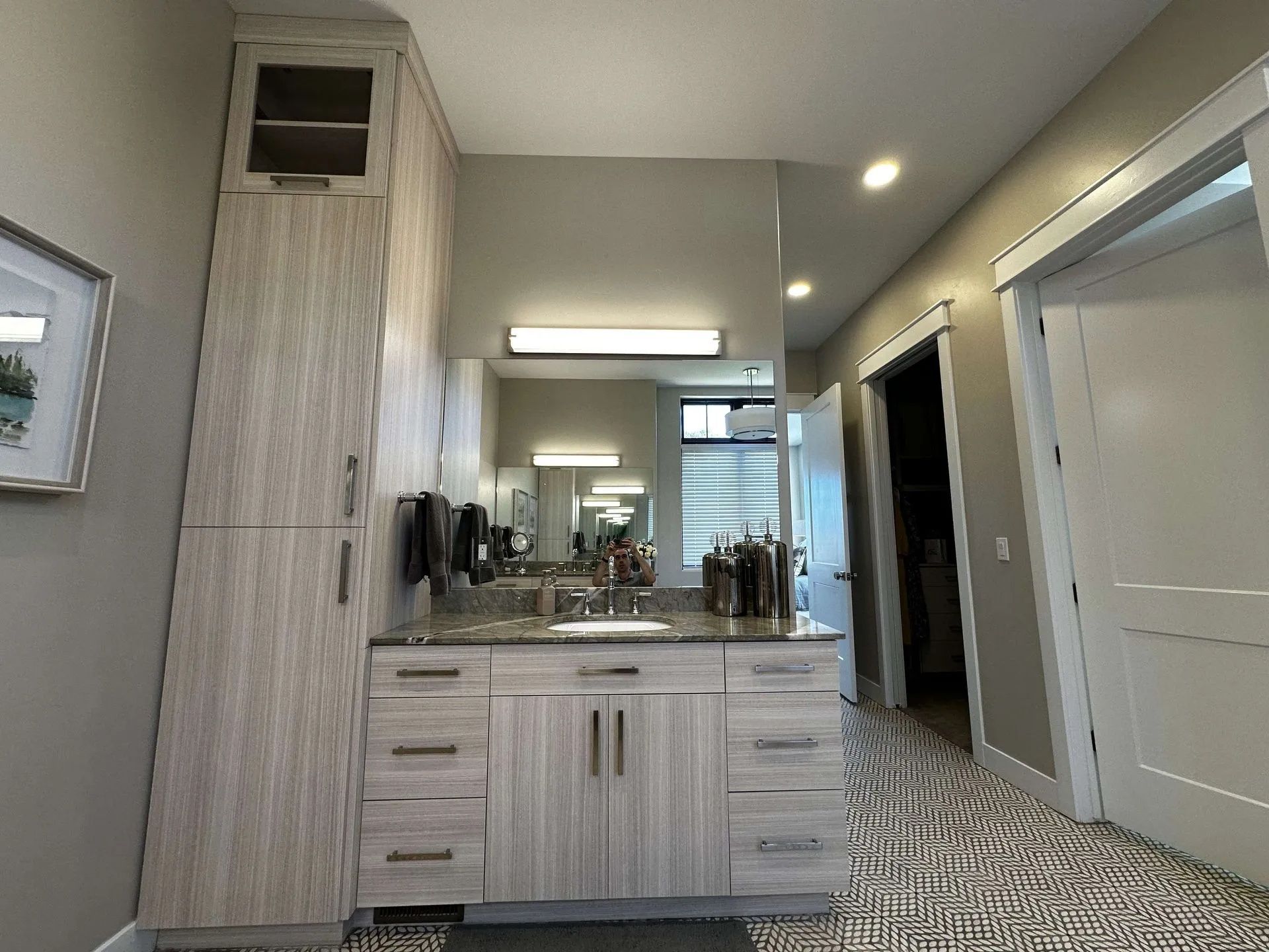 Bathroom with a vanity, mirror, and tall storage cabinet in light wood tones. A hallway with closed doors is on the right.
