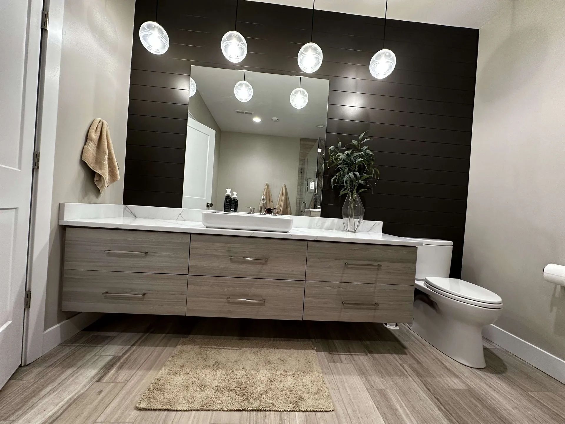 Modern bathroom with floating vanity, toilet, mirror, decorative wall, pendant lights, and wooden floor.