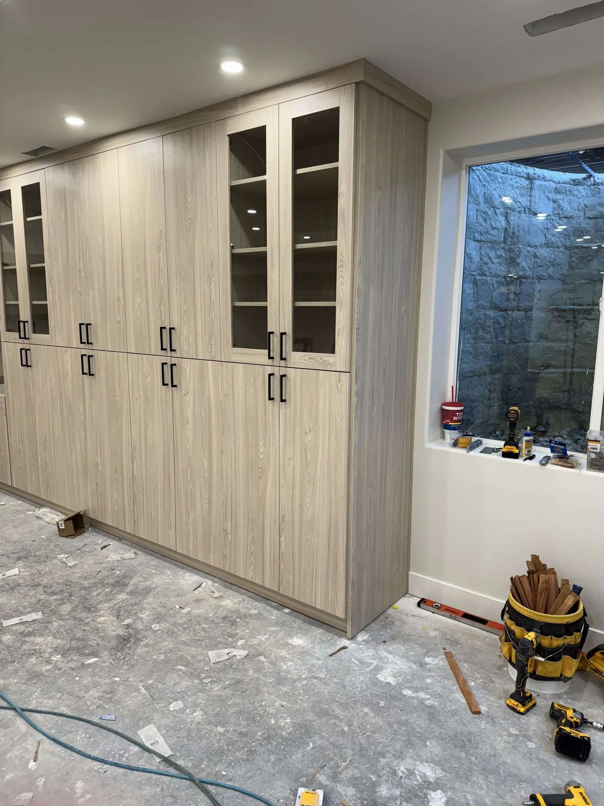 Cabinets under construction next to a window. Light wood grain cabinets, glass-front upper cabinets, tools on the floor.