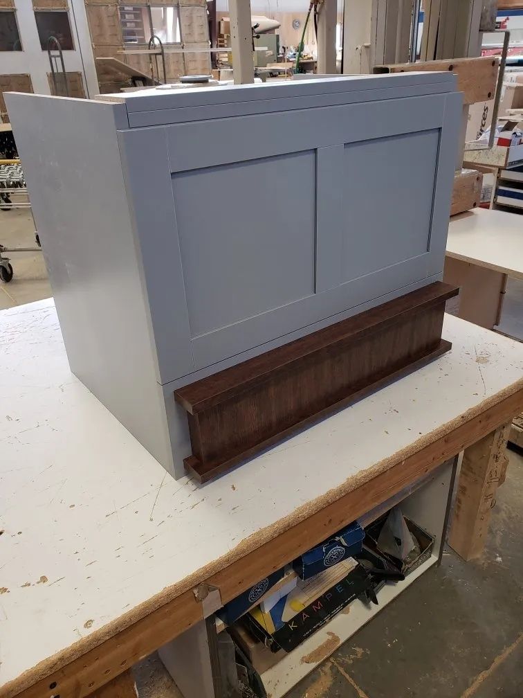 Gray painted wooden cabinet with brown trim sitting on a white workbench in a workshop.