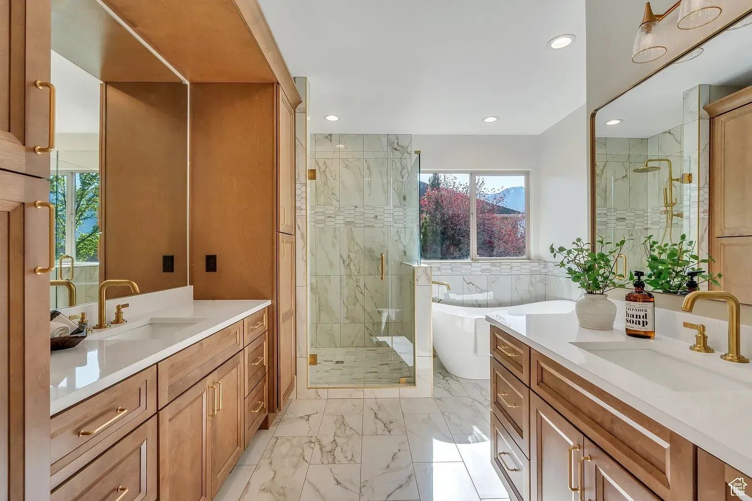 Spacious, bright bathroom with double sinks, glass shower, and soaking tub; light wood cabinetry and marble tile.