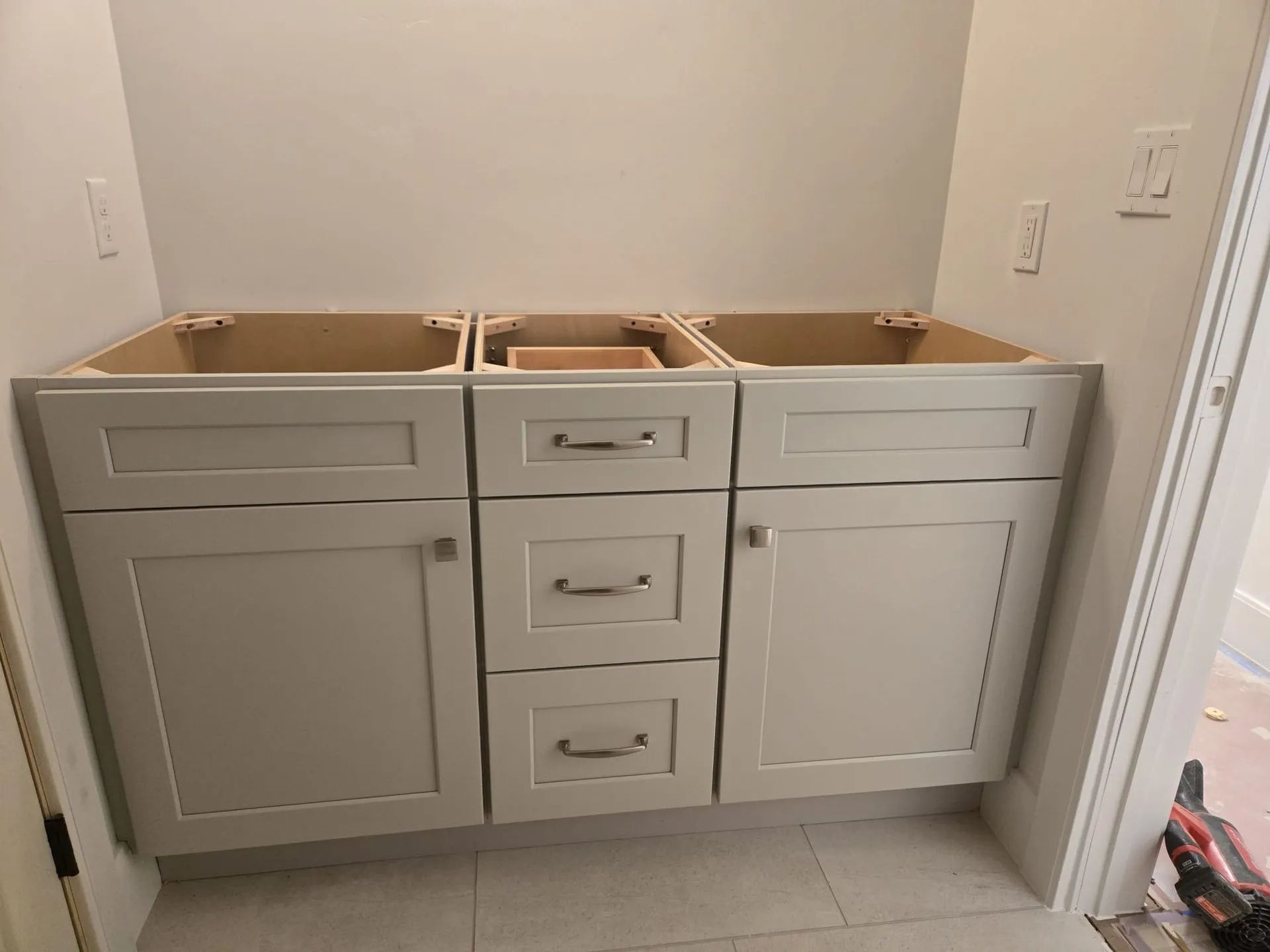 Bathroom vanity cabinets, light gray, installed against a wall, with unfinished interiors and three drawers.
