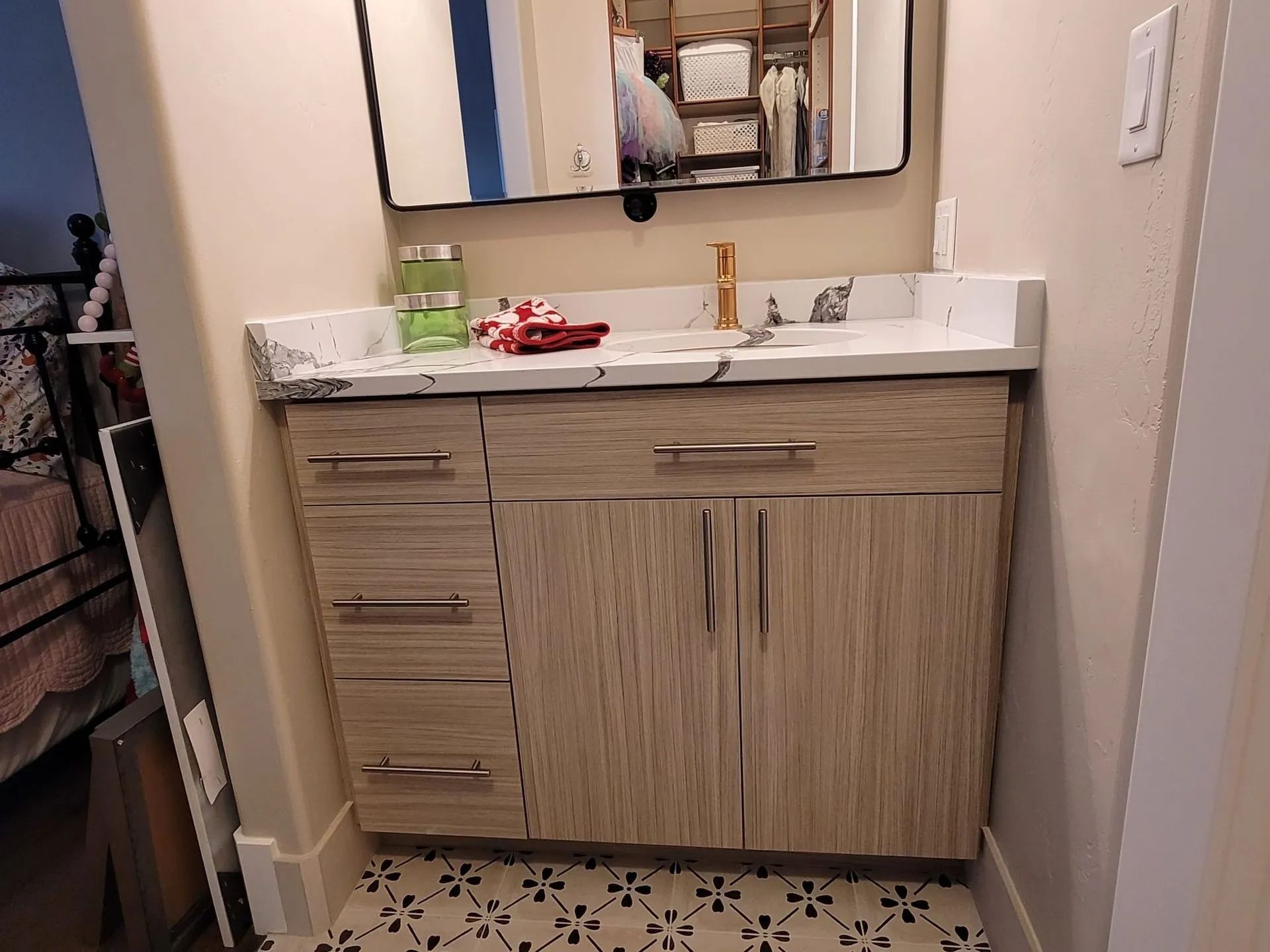 Bathroom vanity with light woodgrain cabinet, marble countertop, and mirror. Black and white patterned tile floor.