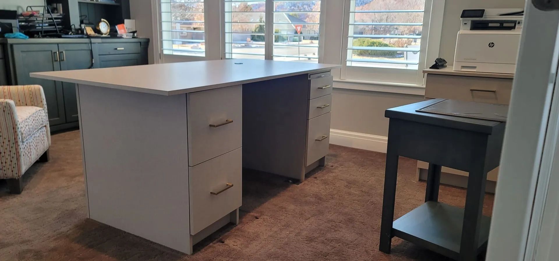 A large white desk with three drawers. A black side table sits beside it near a window.