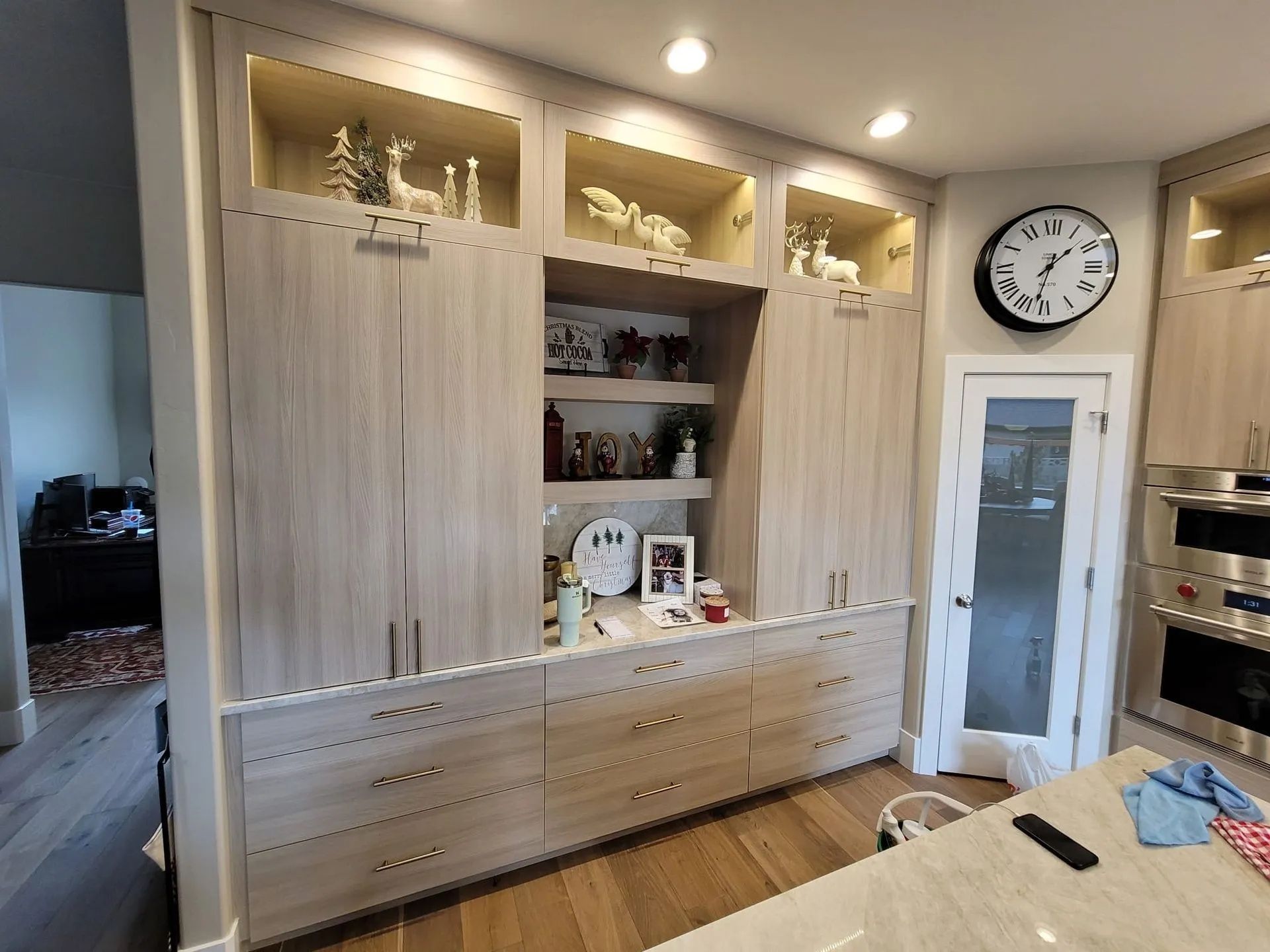 Kitchen cabinets with illuminated shelves, light wood finish, clock on wall, oven, and entryway.