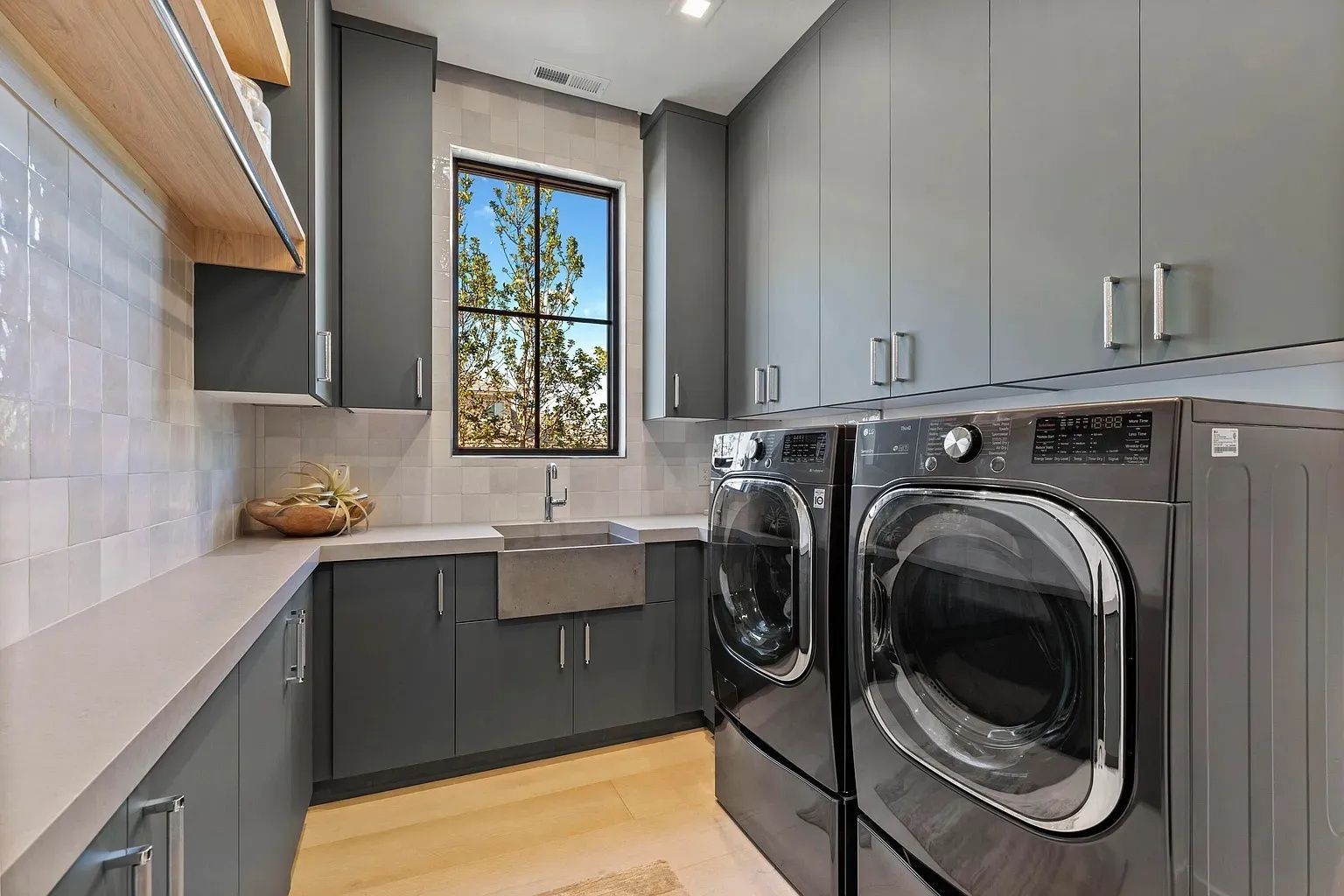 Laundry room with gray cabinets, washer/dryer, sink, window with outdoor view, and light wood shelves.