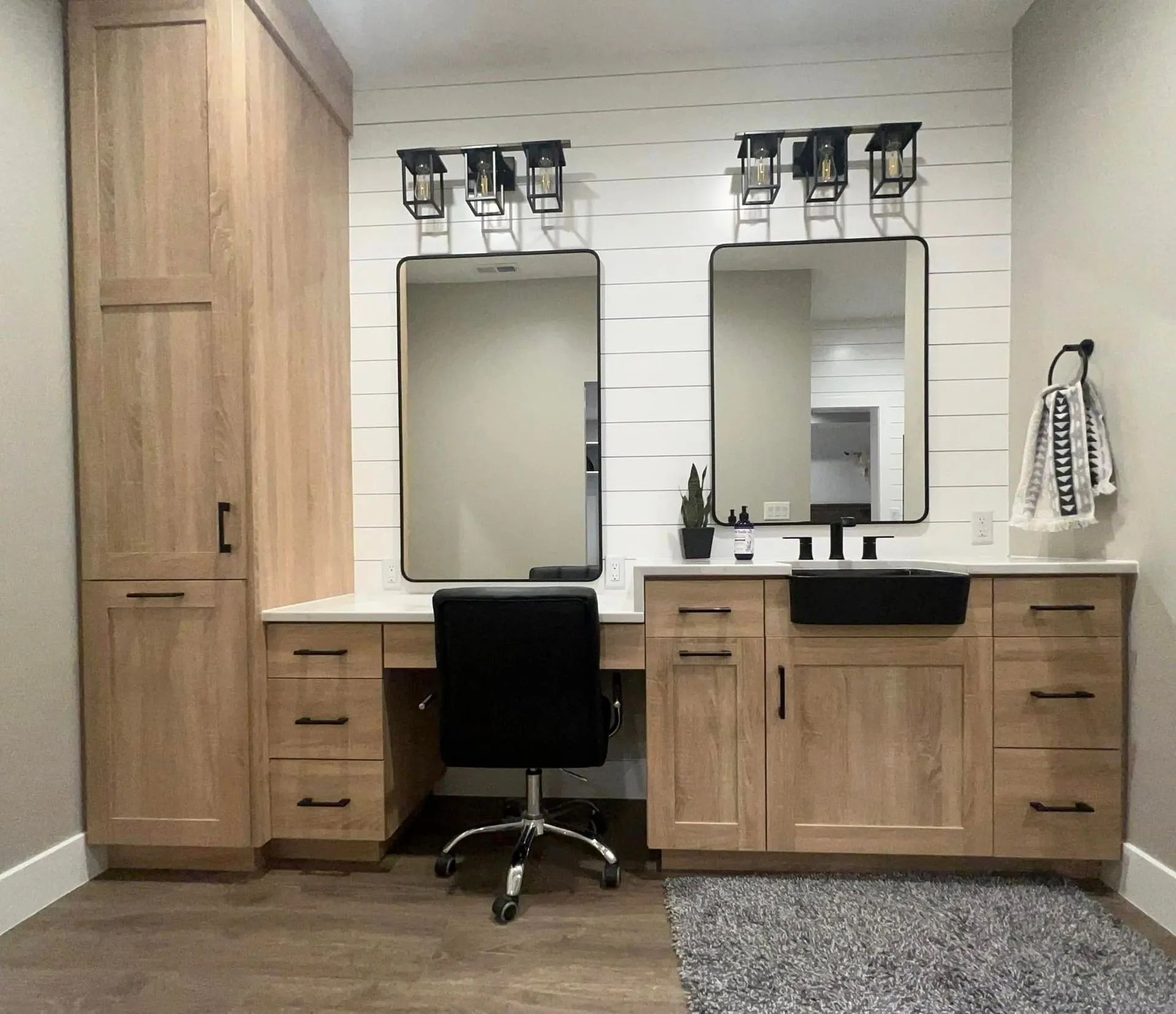 Bathroom with two mirrors, vanities, and a desk. Light wood cabinets, black fixtures, and white wall.