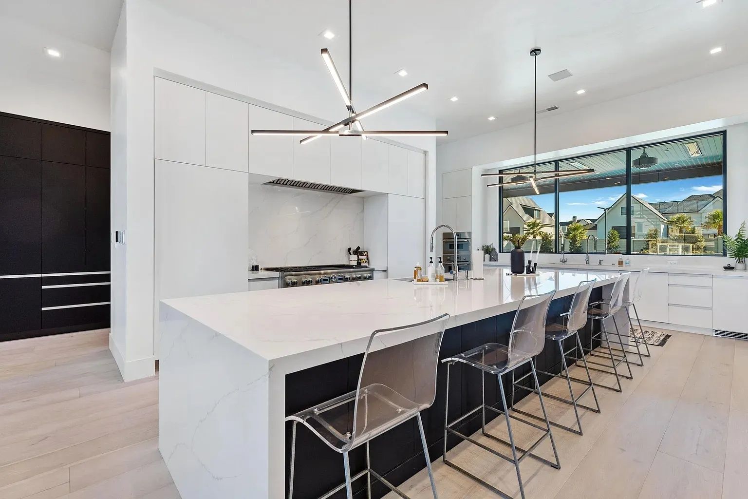 Modern white kitchen with island, seating, and window overlooking a yard. Black cabinetry on the left.