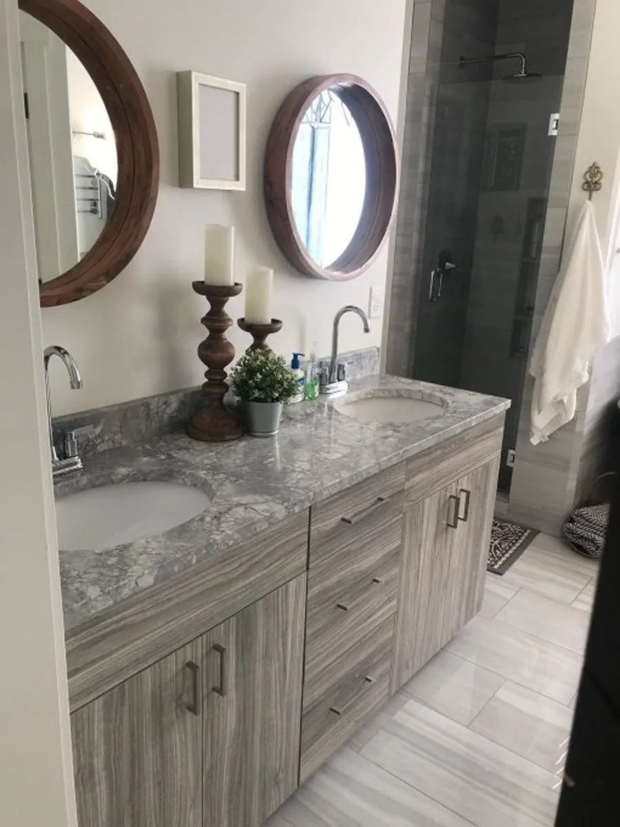 Bathroom with double sinks, gray cabinets, marble-like countertop, mirrors, and a glass shower.