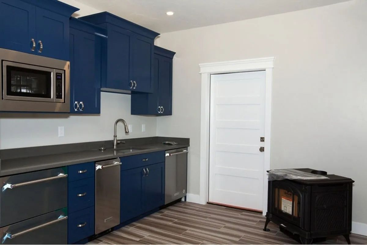Blue kitchen cabinets with stainless steel appliances, sink, and a white door on the right. A wood-burning stove sits on the floor.