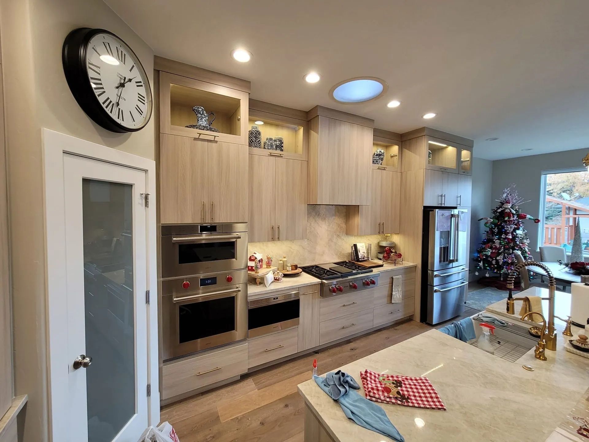 Modern kitchen with light wood cabinets, stainless steel appliances, and a Christmas tree in the background.