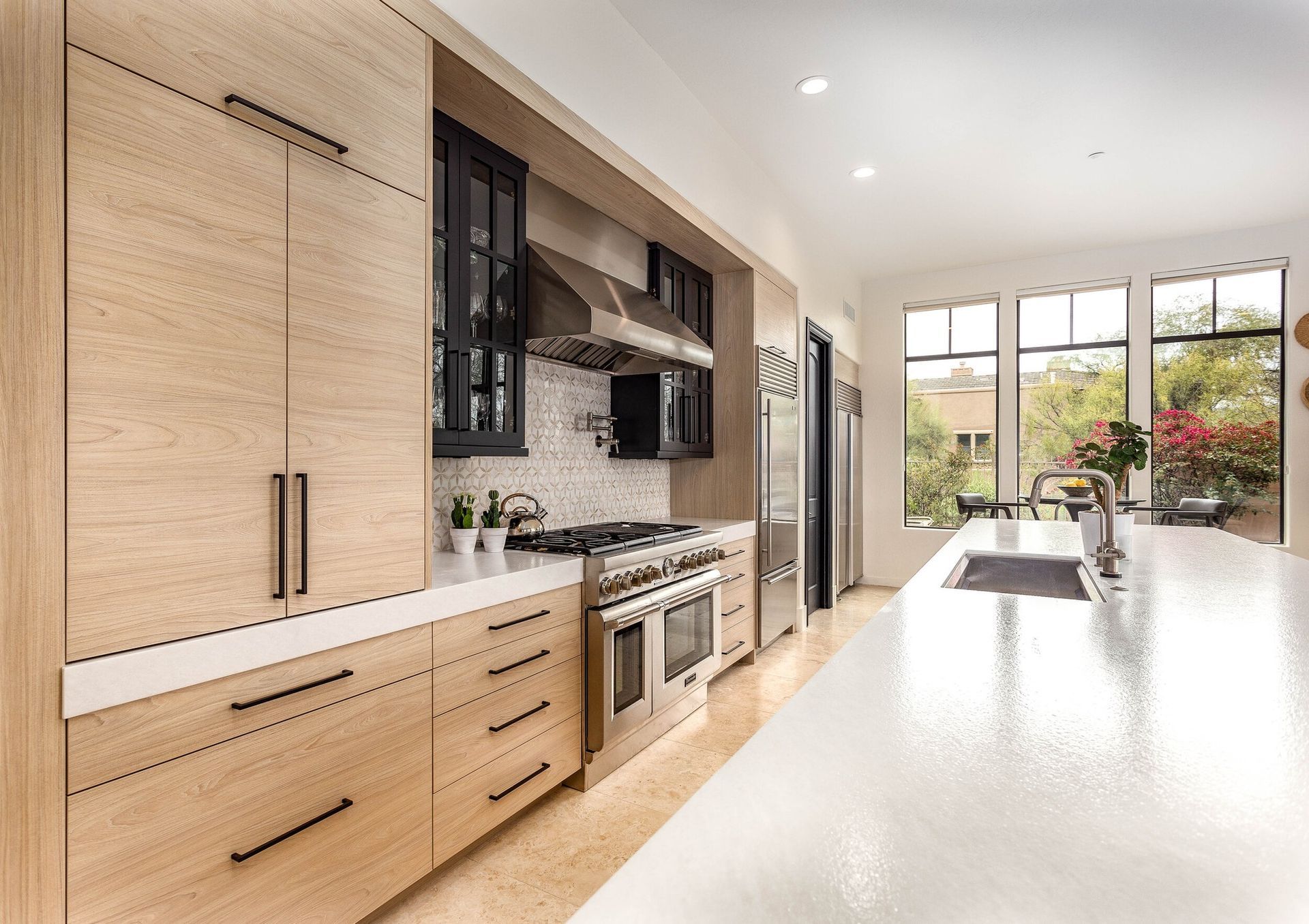 Modern kitchen with light wood cabinetry, white countertops, stainless steel appliances, and large windows.