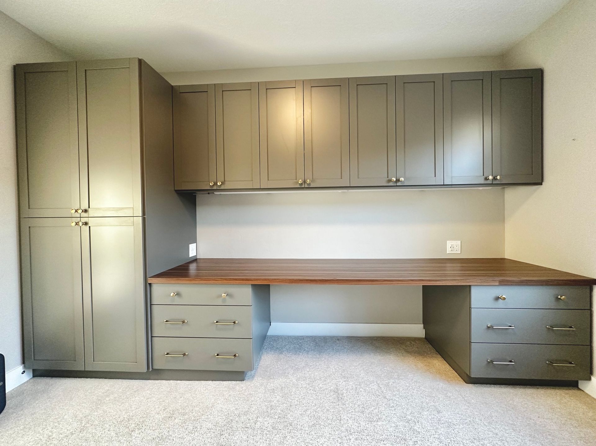 Built-in home office with tall cabinet, overhead storage, desk with drawers, and wood-grain desktop; gray cabinets.