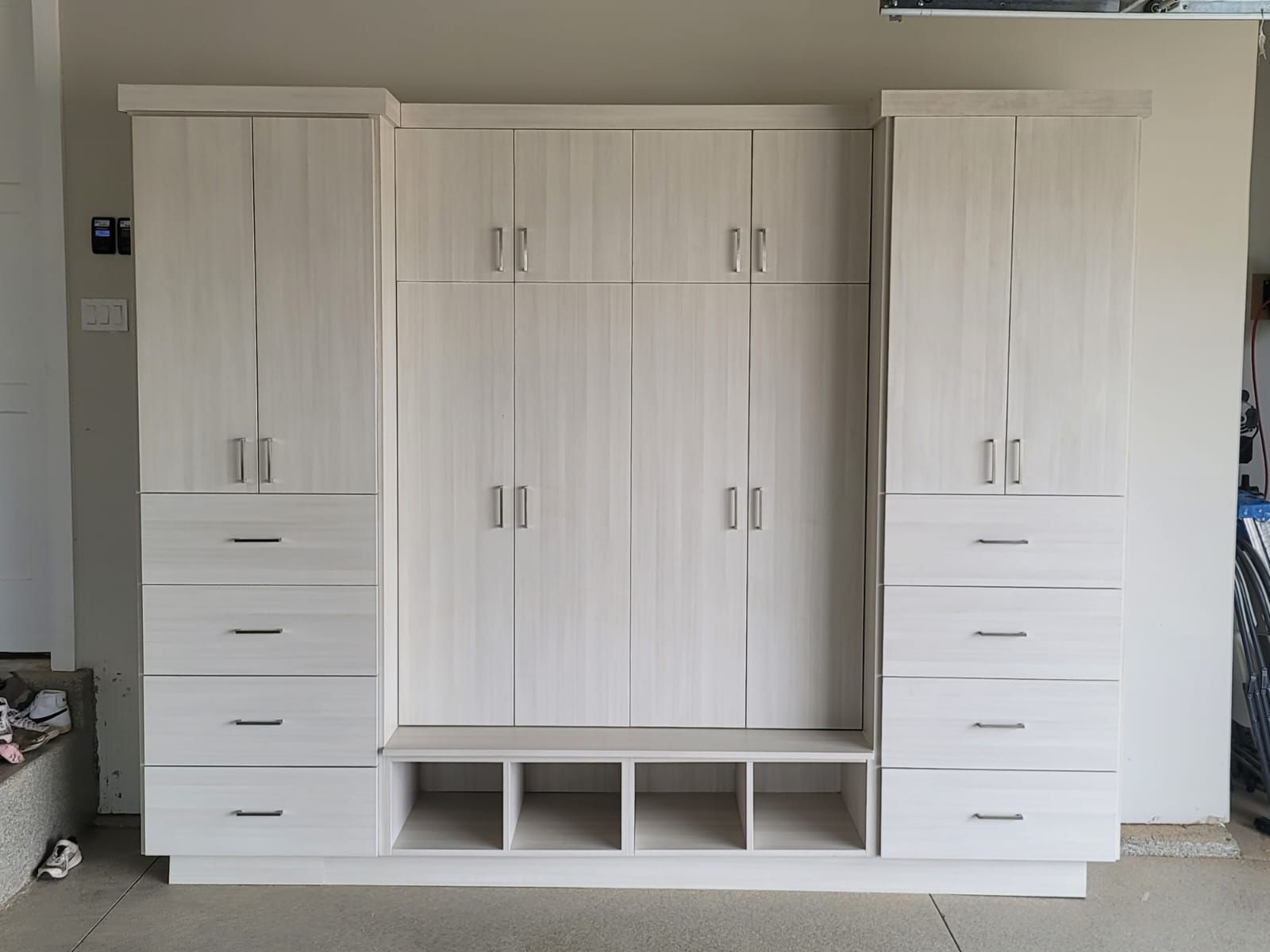 Built-in, light-colored wooden storage unit in a garage, with cabinets, drawers, and bench seating.