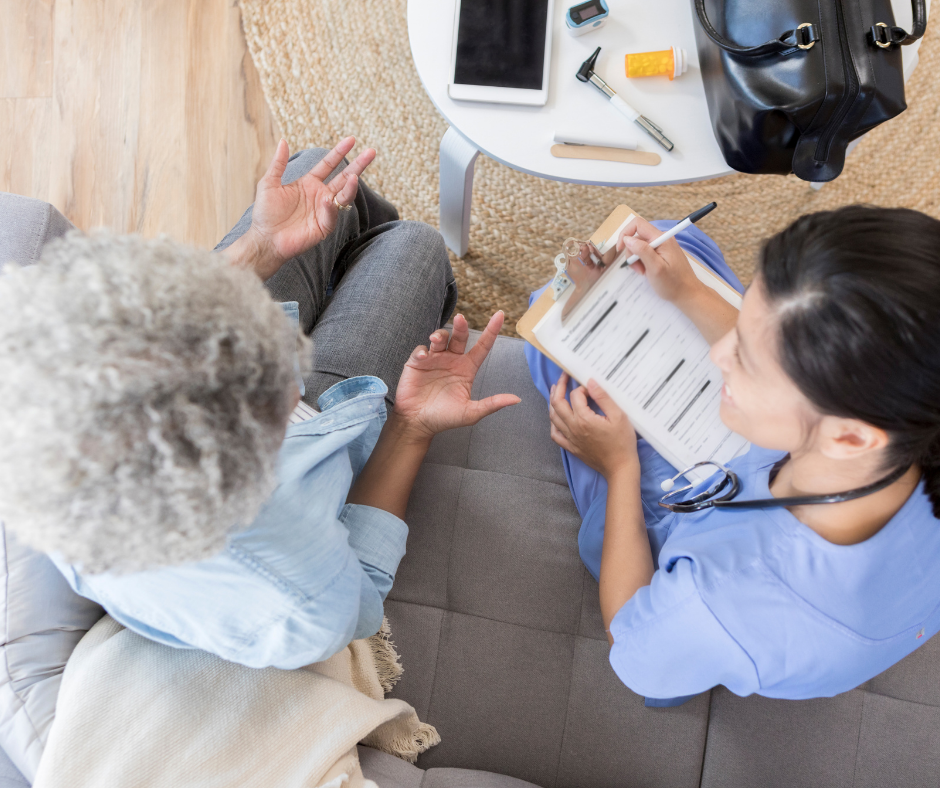 A nurse is writing on a clipboard while talking to an elderly woman on a couch.