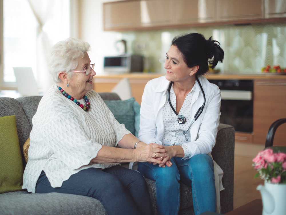 An elderly woman is sitting on a couch holding hands with a doctor.