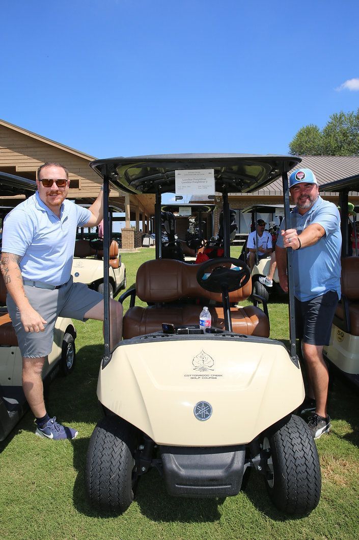Two men are standing next to a golf cart on a lush green field.