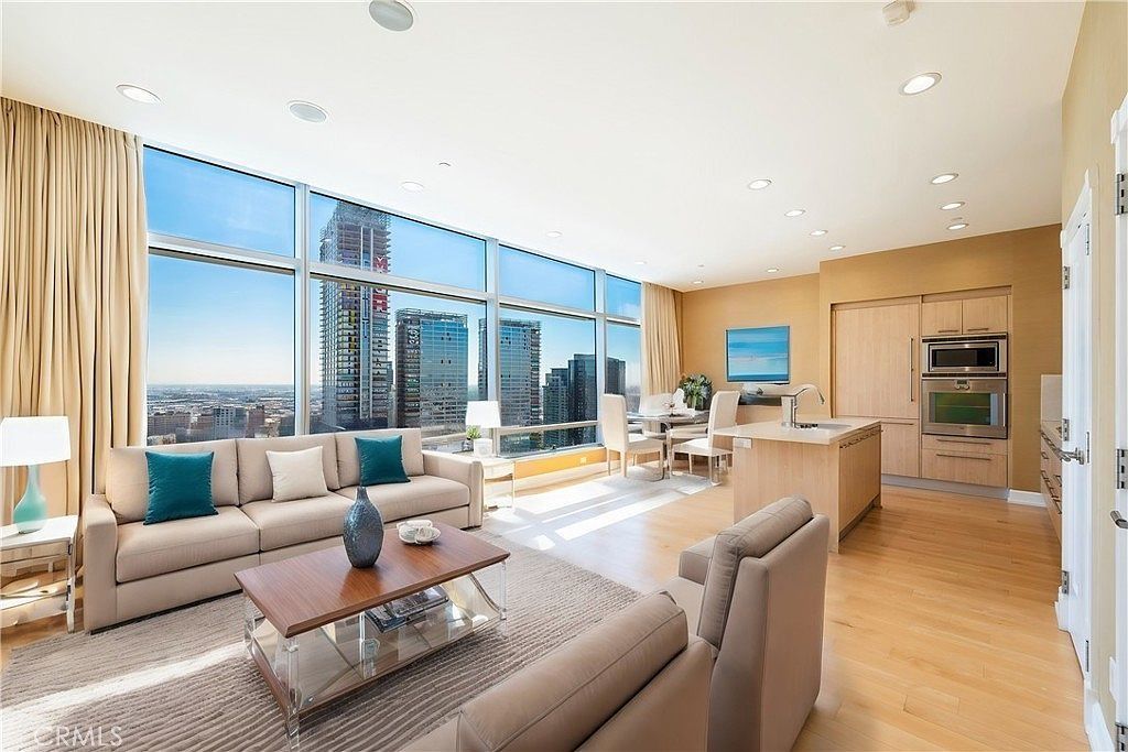 Bright modern living room with floor-to-ceiling windows, beige sofas, and an open kitchen with city views