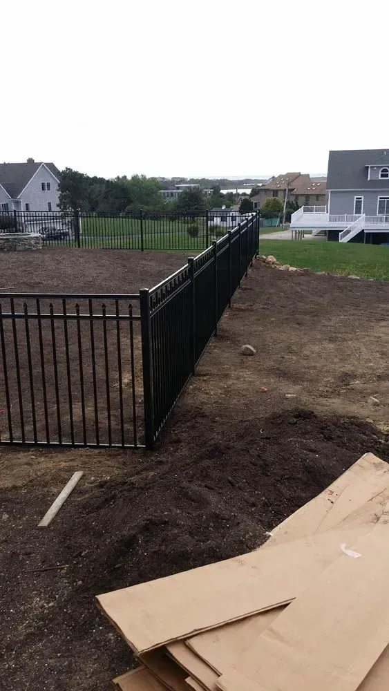 Black metal fence bordering a yard filled with dark soil, with houses and trees in the background.