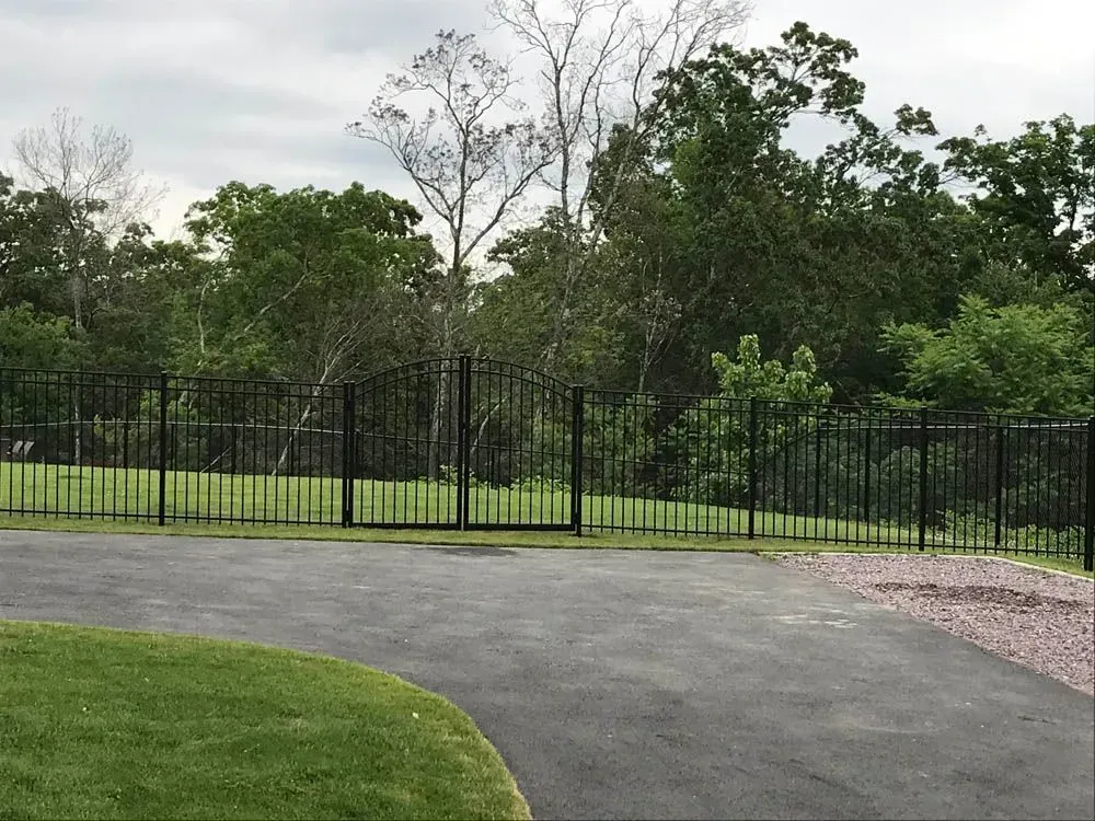 Black metal fence encloses a green lawn with trees in the background, asphalt driveway in foreground.