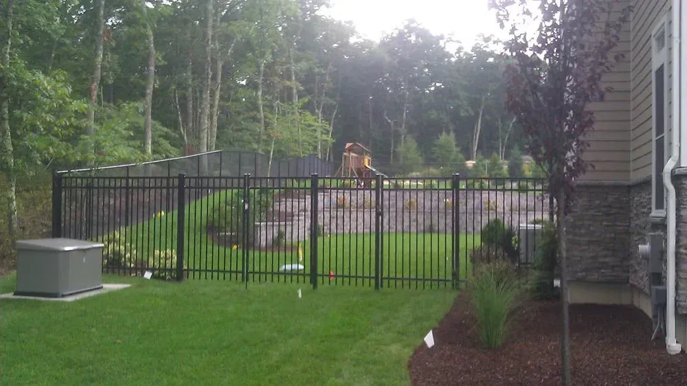 Black fence encloses a yard with green grass and landscaping, next to a house with stone facade.