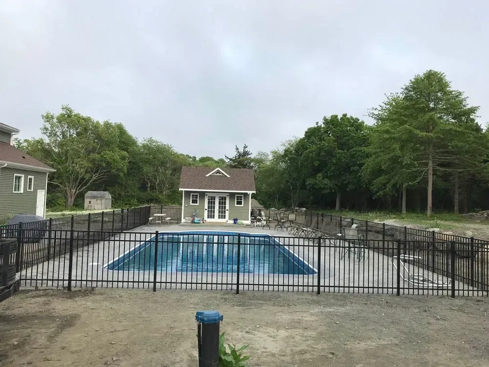 A rectangular pool enclosed by a black fence, small building, and trees on a cloudy day.