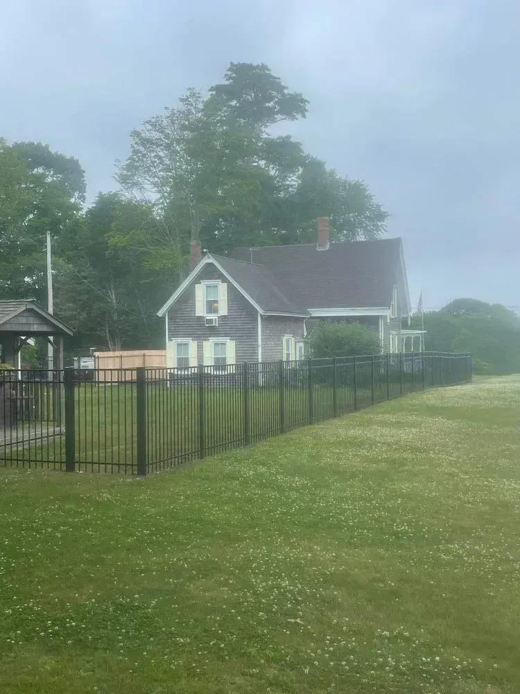 Gray house with black fence in front, green lawn, overcast sky.