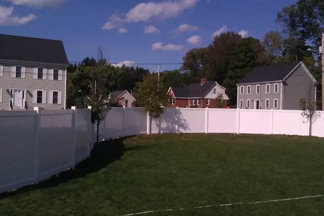 White fence encloses a green yard in a residential area, with houses and a blue sky visible.