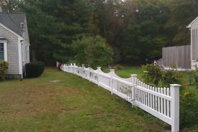 White picket fence curves along a grassy yard between two houses, with a tree line in the background.