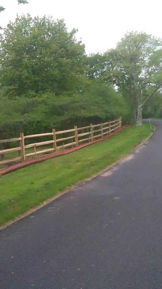 Wooden split-rail fence bordering a green lawn and a paved road next to a line of green trees.