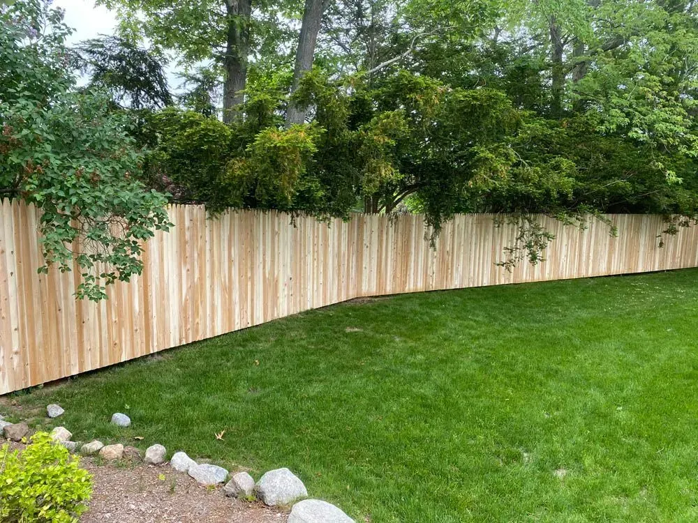 Wooden fence curves along a green lawn, edged with rocks, trees in the background.