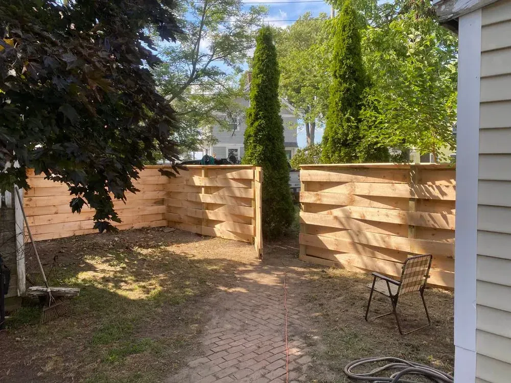 Wooden fence in a yard with a brick pathway. Two tall green trees stand beyond the fence.