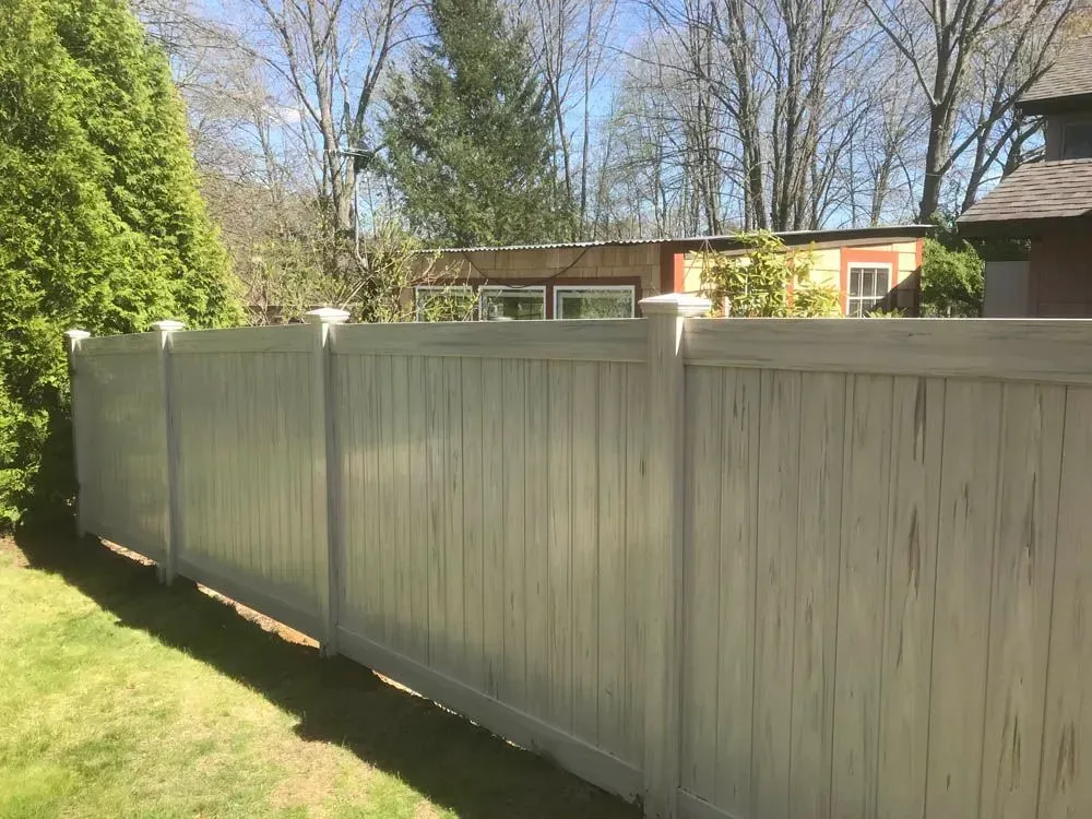 White vinyl fence in a backyard, green grass, trees, and a house in the background.