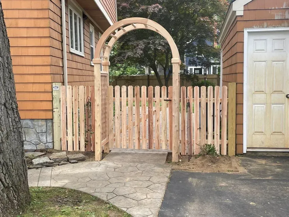 Wooden picket fence with arched gate and adjacent garage door.