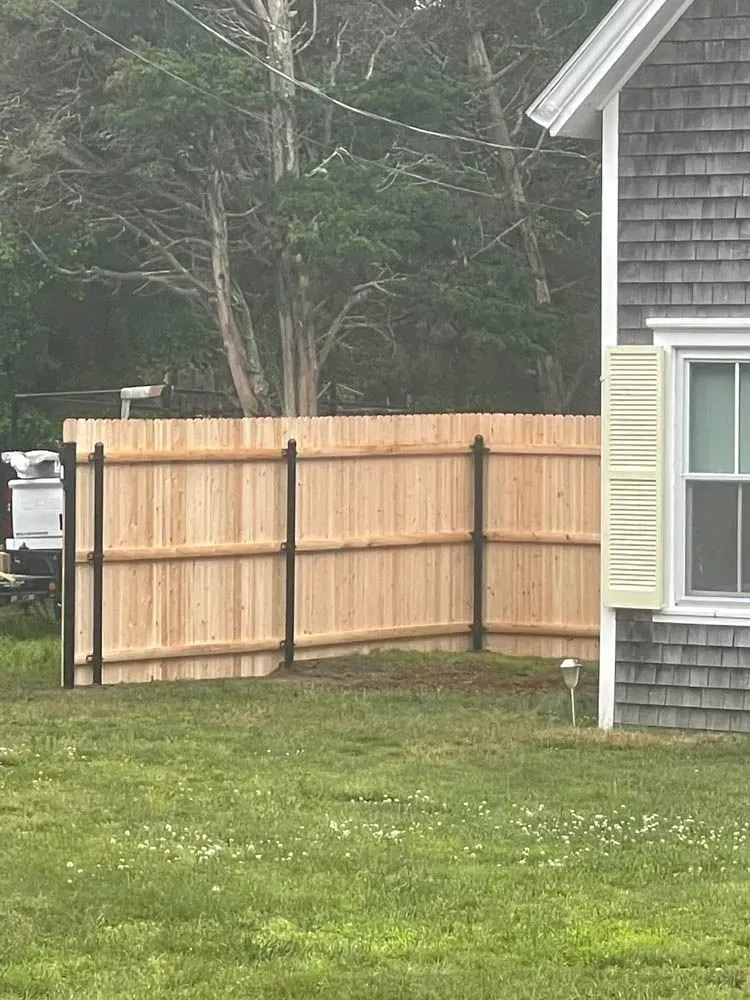 A wooden privacy fence, with black posts, in front of a gray shingled house. Green grass in the foreground.