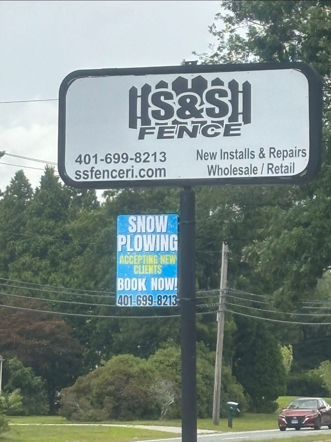 Hastings Fence roadside sign with phone number, next to a blue snowplowing sign by a grassy roadside