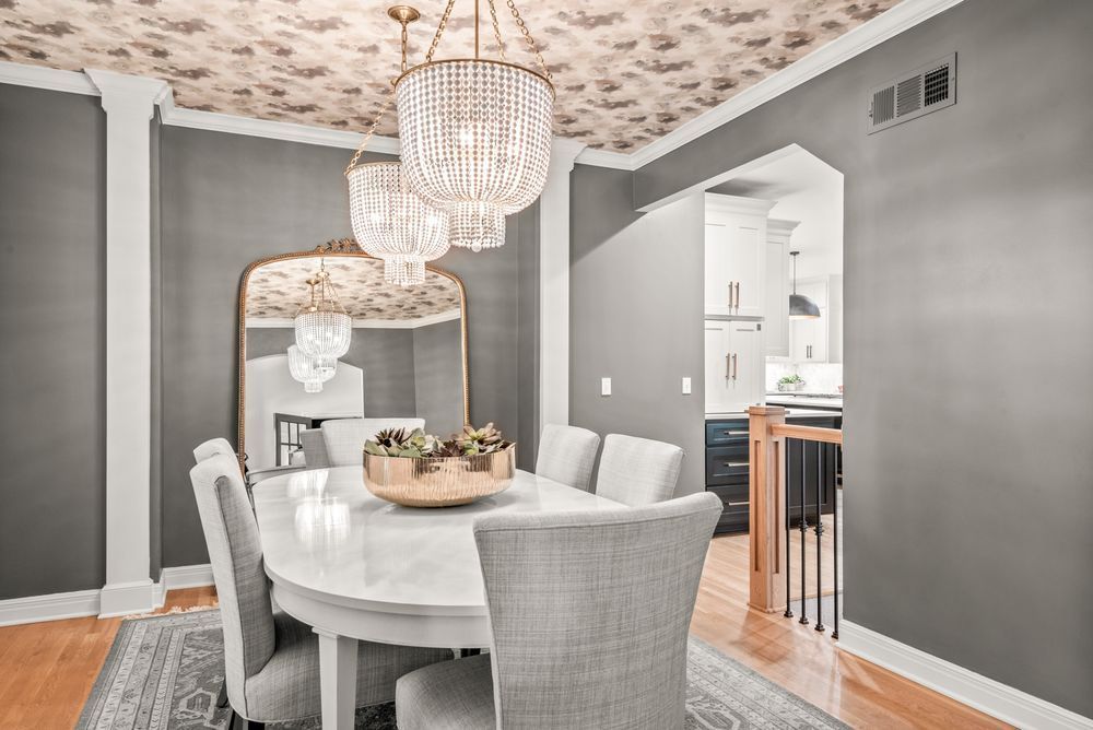 Elegant dining room with a white oval table, grey chairs, a large gold-framed mirror, and crystal chandeliers. Gray walls and a floral patterned ceiling.