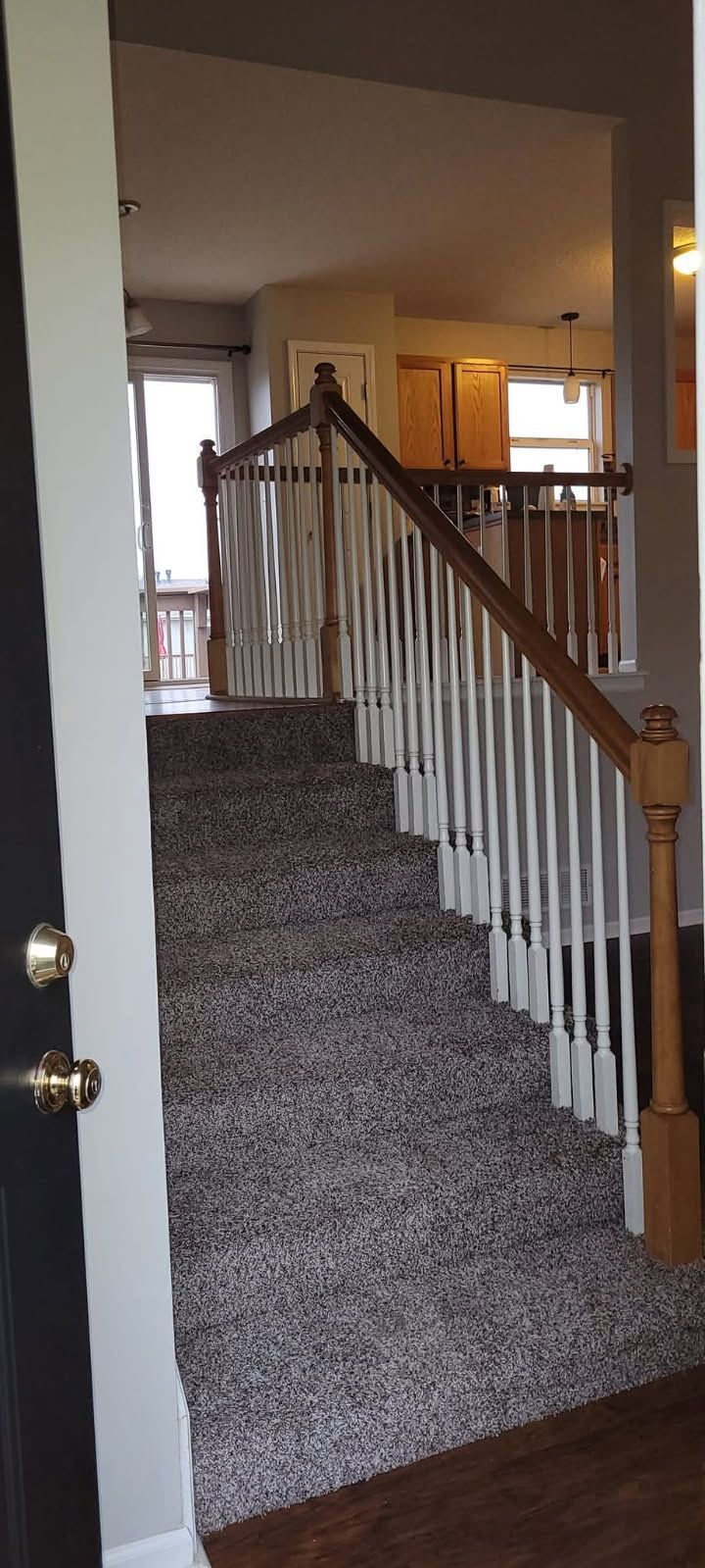 A view from a doorway shows stairs with white balusters, a wooden banister, and gray carpeting. The kitchen is visible in the background.